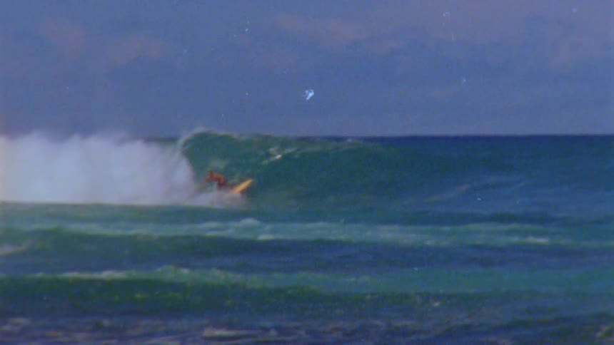 Man rides a surfboard over a big wave near the coast caught on vintage film
