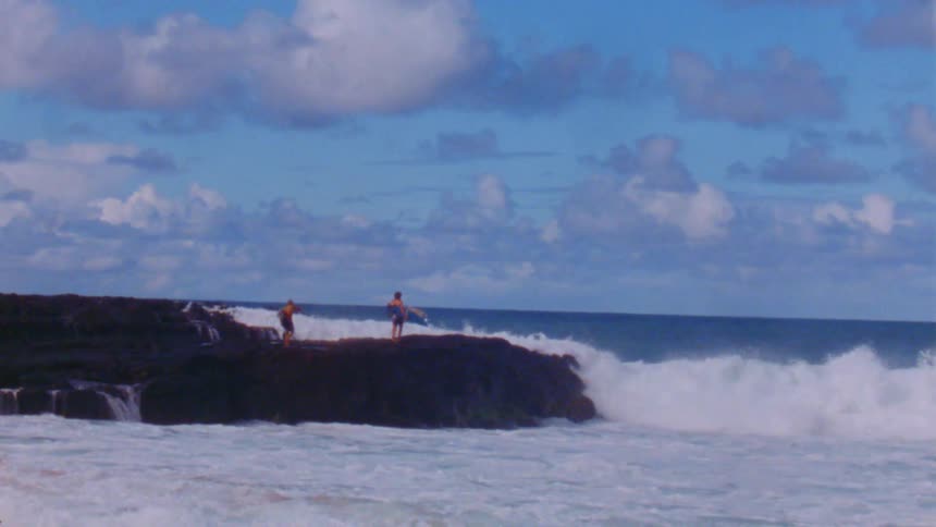 Three boys are standing on the rocks holding surfboards and the waves splash as they crash into the rocks