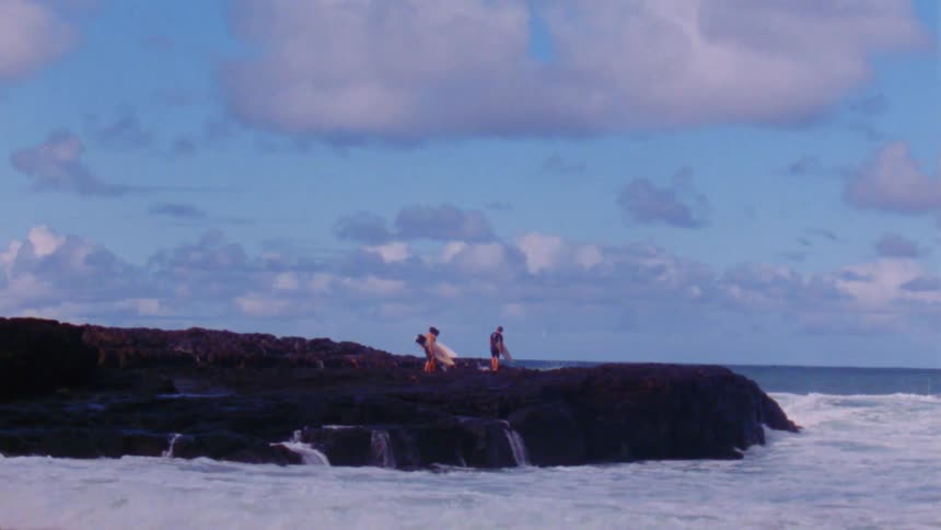 Surfers walk along rock coast carrying surfboards as water flows down rock into ocean