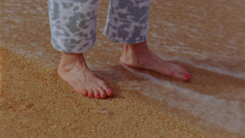 Close up of the feet of a barefoot person standing on the sand of a beach caught on vintage film