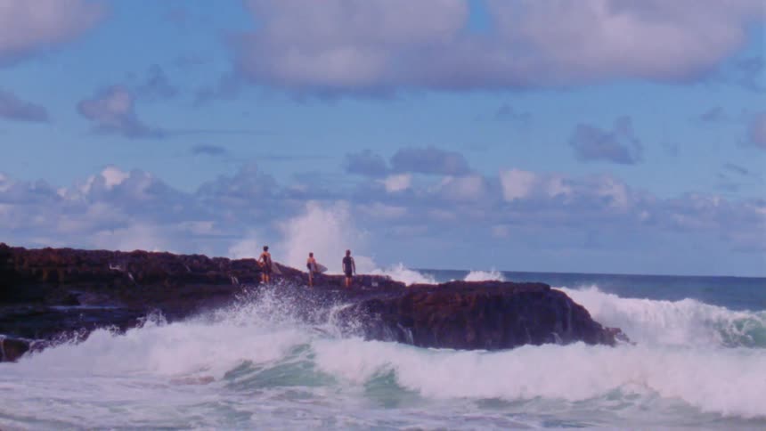 Waves crashing against rocks on the coastline while three people with surfboards stand on the rocks