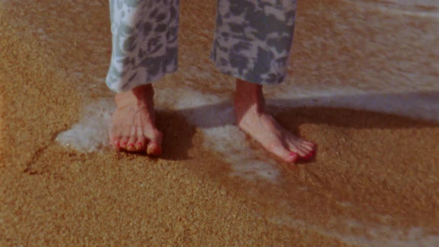 Woman in white gray pants stands barefoot with red toenails on the sand beach near the ocean