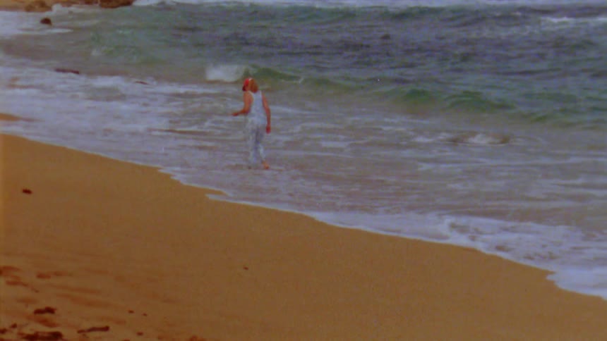 A woman wearing a red hat and overalls is walking along the beach as waves roll into shore