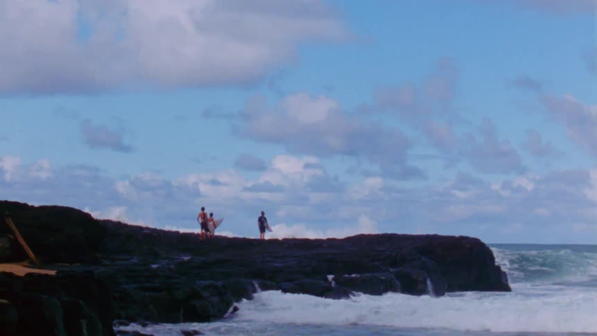 Male surfers stand holding surfboard staring at ocean as waves crash and spray