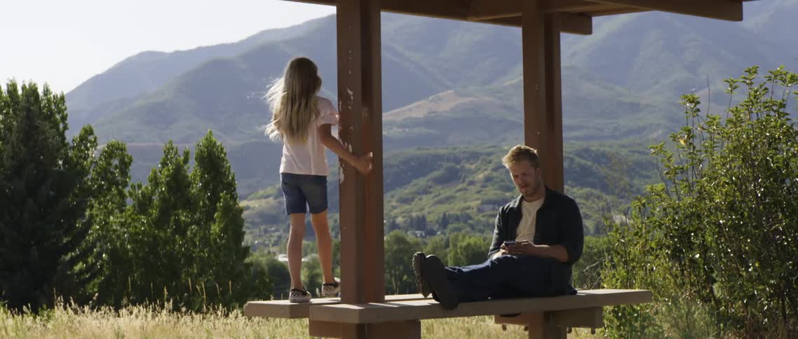 A man is stretched out sitting on a bench using his cell phone as his daughter walks around on the bench and there are mountains in the distance