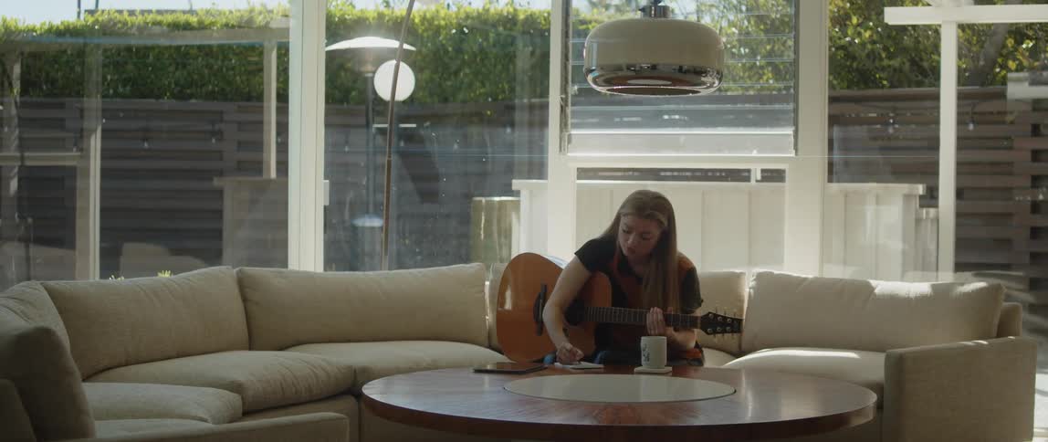 A female is sitting on a couch with a guitar and writing something on a notepad