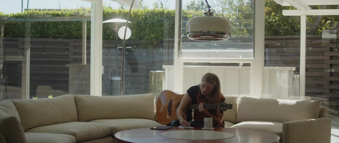 Woman with brown hair black and orange shirt plays acoustic guitar and takes notes sitting on beige couch in the living room