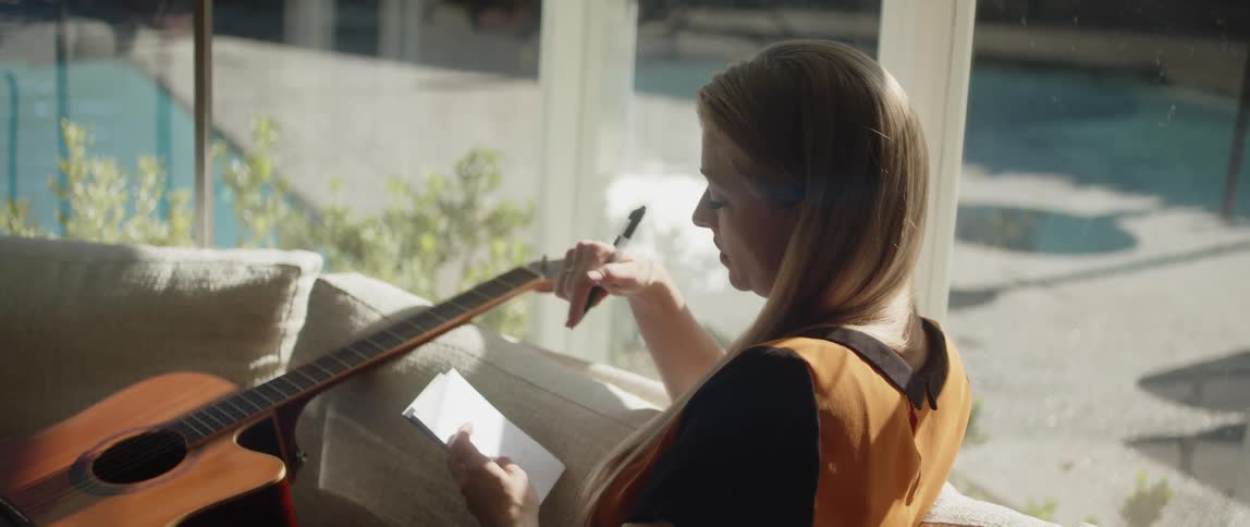 A woman is sitting on her couch near windows holding a notepad and writing with an acoustic guitar sitting next to her