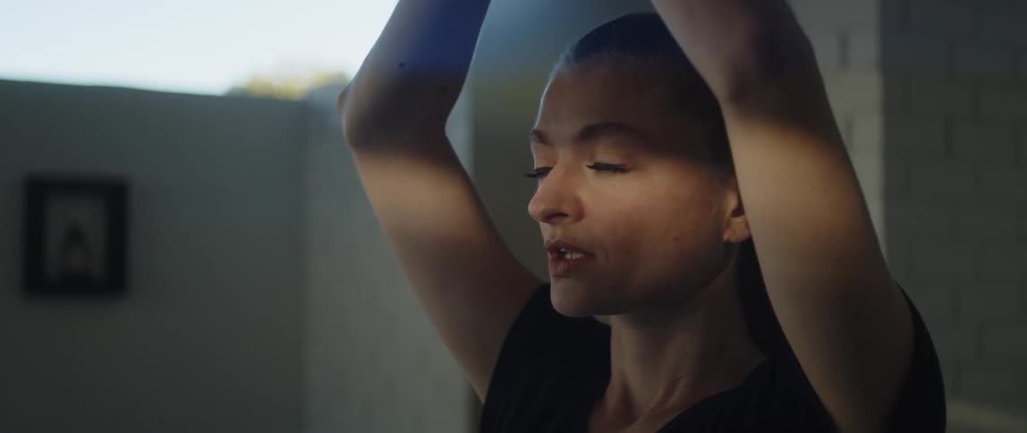 Light shines on a womans face as she raises her arms over her head and moves them in front of her face as she practices yoga