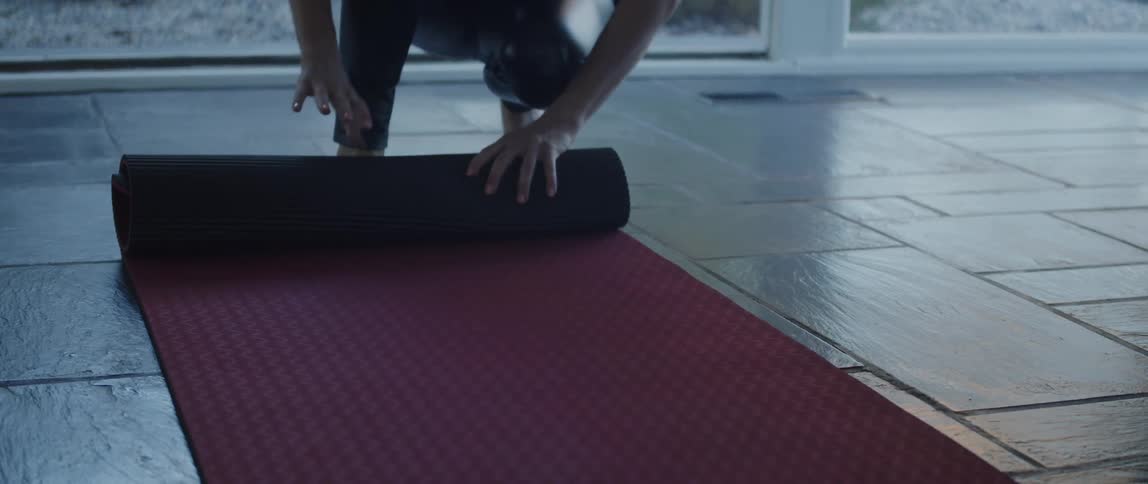 A person kneeling down rolling up a red yoga mat and then standing up
