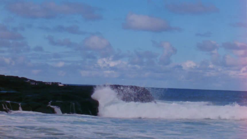 Waves crash onto beach and elevated stone shore spraying foam under a cloudy sky