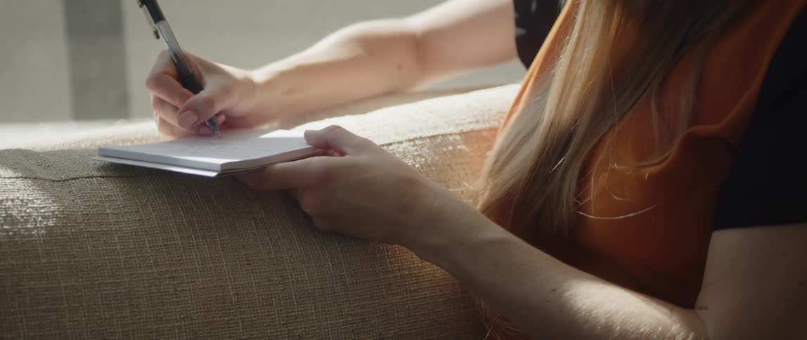 A woman writes on a notepad while sitting on a sofa indoors during the day