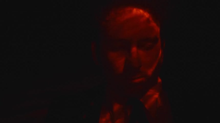 A man dressed in a suit is standing under water with his eyes closed and barely illuminated by a red light