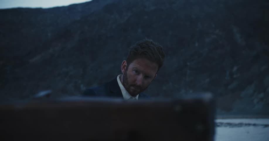 Bearded man sitting in the desert wearing a business suit turns to look at a suitcase