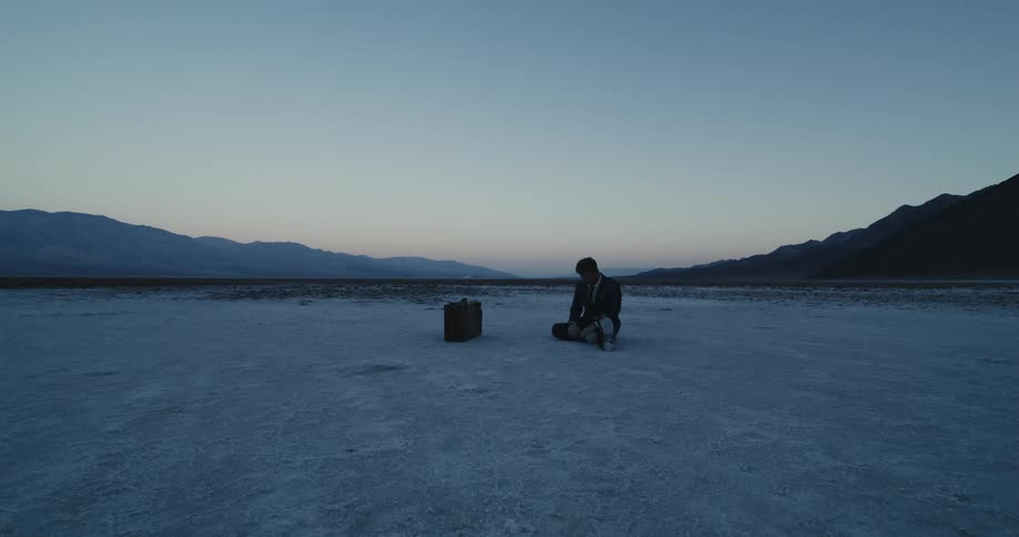 A man in a suit is sitting on the snow covered ground and there is a briefcase near him and mountains in the distance