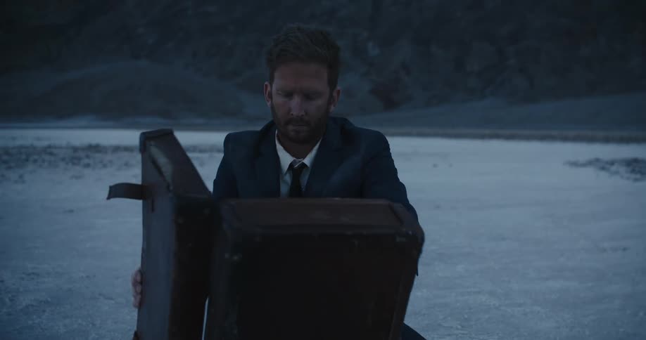 Bearded man sitting in the desert wearing a business suit turns a suitcase around