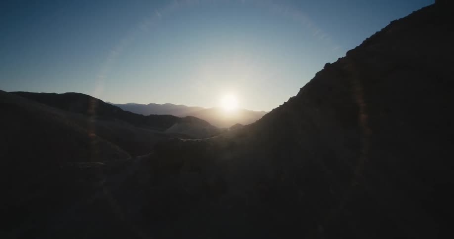 The sun shines brightly on the horizon illuminating mountains under a clear sky