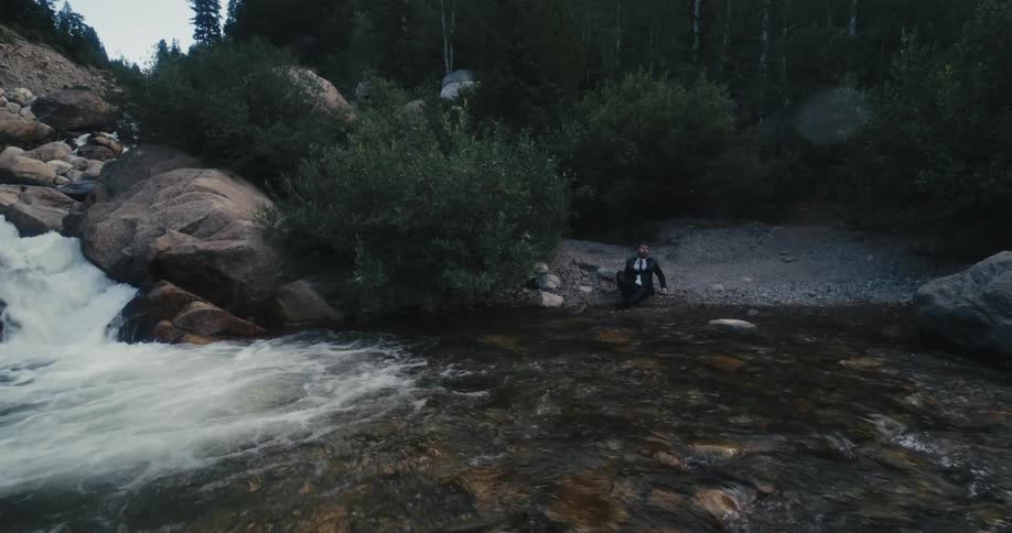 A man in a suit suddenly emerges from beneath the waters of a river and struggles to walk his way to the shore