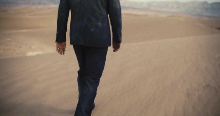 A man wearing a dirty suit is walking in the desert and there are mountains in the distance