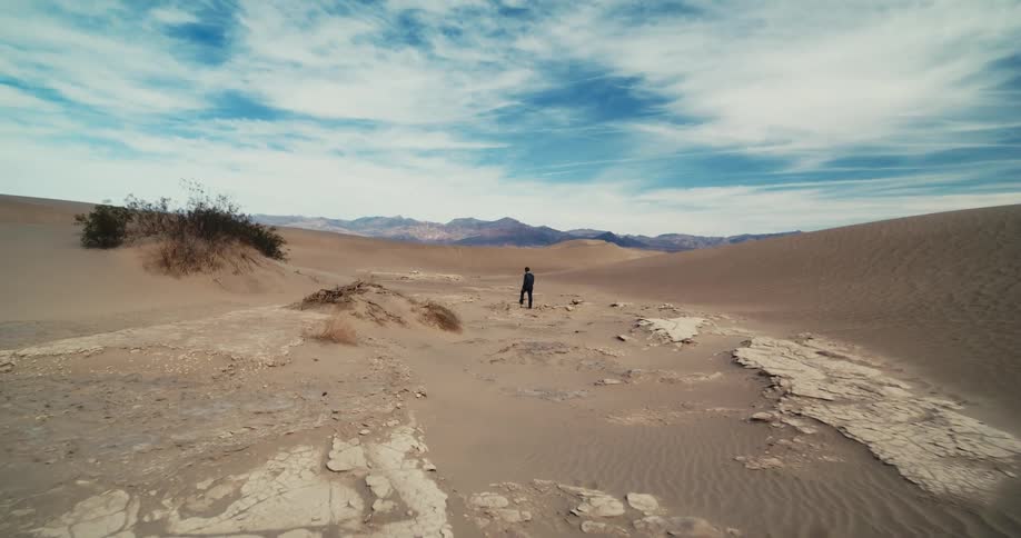 A man wearing a suit is walking in the desert among rocks and dunes with mountains in the distance under a cloudy sky