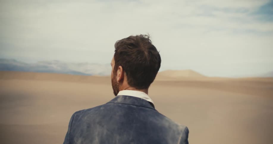 A man wearing a dirty suit is standing in the desert looking around and there are mountains in the distance