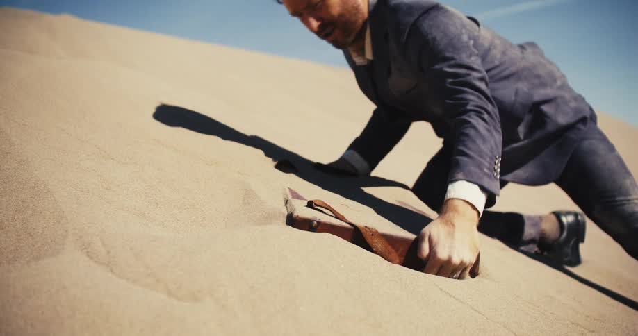 A man is kneeling on a desert hill as he pulls a briefcase out of the sand