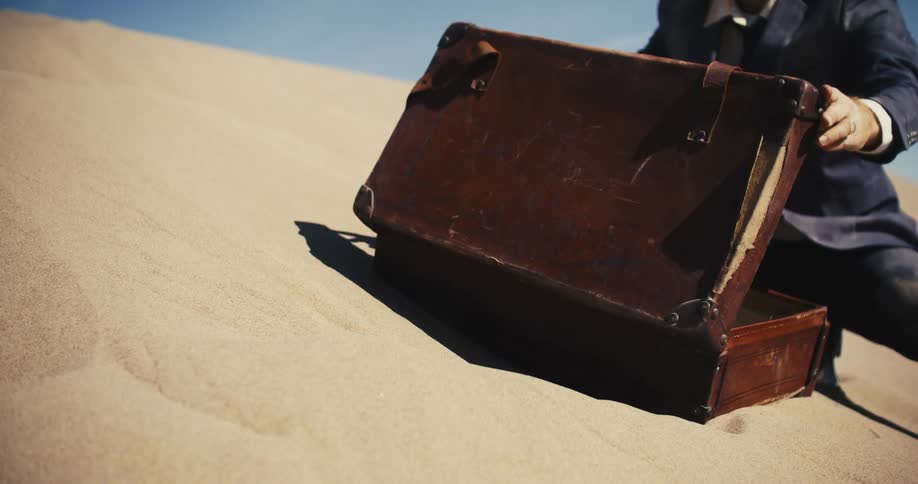 A man wearing a suit is sitting in the desert sand as he opens a briefcase and then closes it