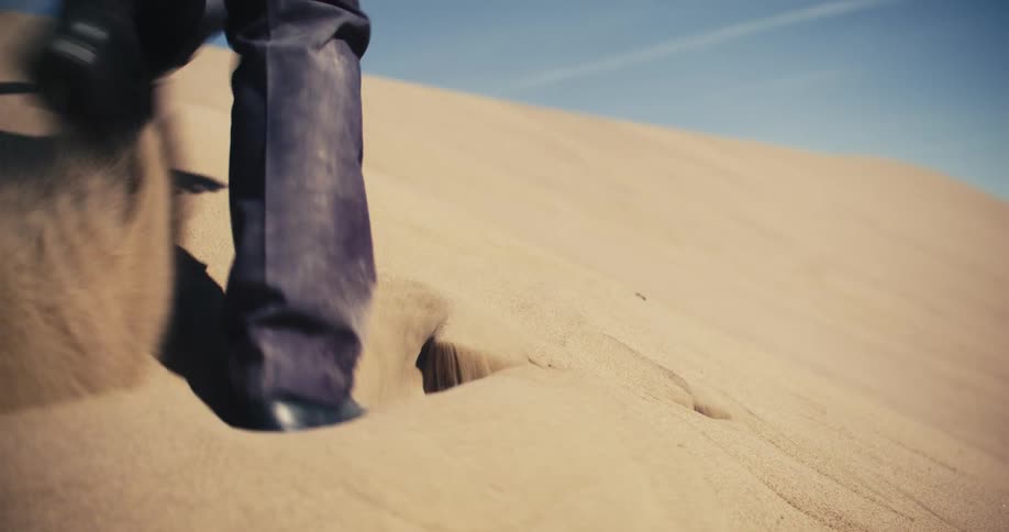 A man wearing a suit is walking in the desert and he finds a briefcase in the dune and digs it out of the sand