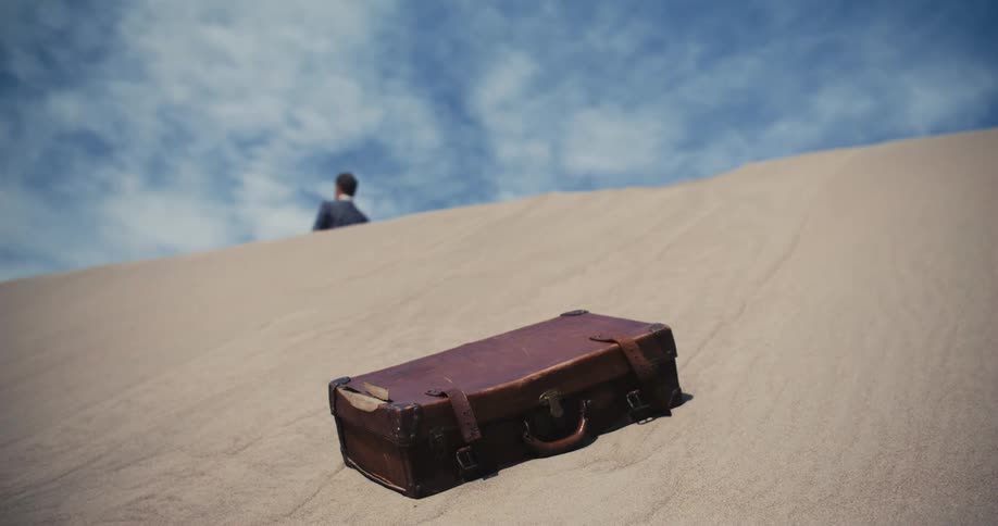 A man in a suit is walking up a desert hill leaving a briefcase in the sand behind him