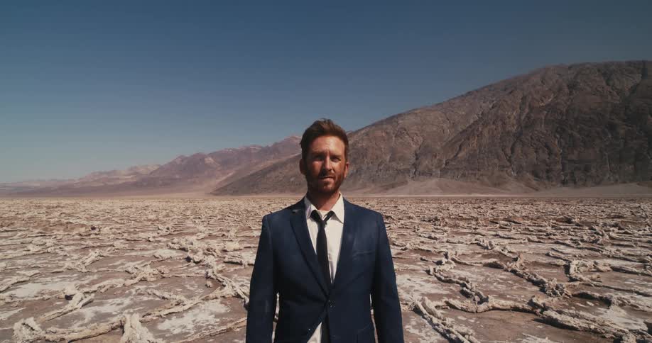 The sun is shining on the face of a man in a suit standing in the desert looking tired and confused