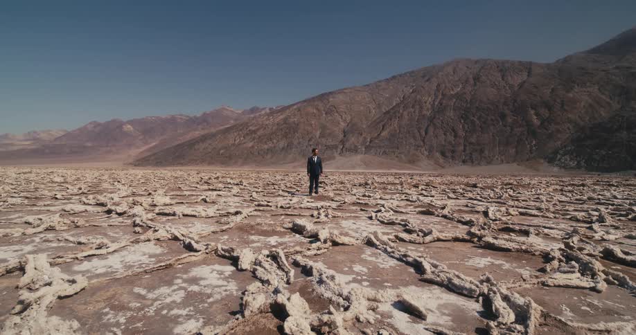 A man wearing a suit is standing in the desert near mountains under a clear sky