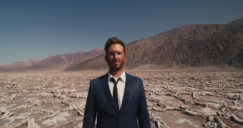 A man wearing a suit is standing in the desert looking around and there are mountains in the distance
