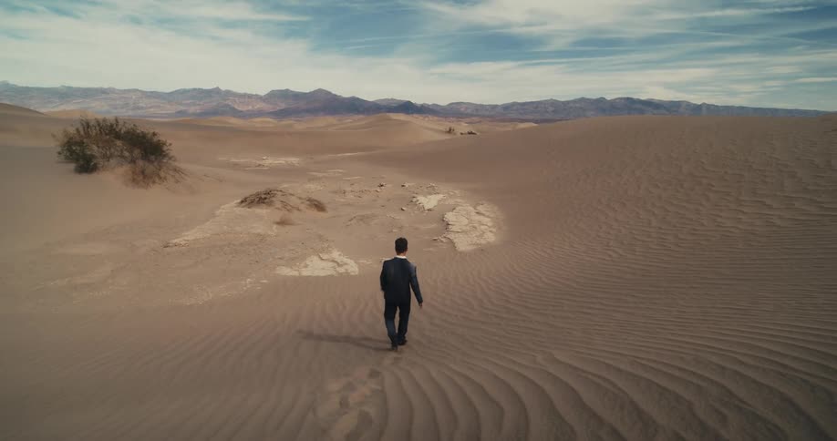A man wearing a suit is walking and stumbling in the desert among rocks and dunes with mountains in the distance under a cloudy sky