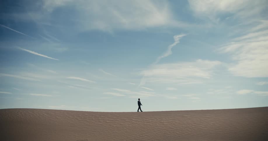 A man is walking across the desert under a cloudy sky
