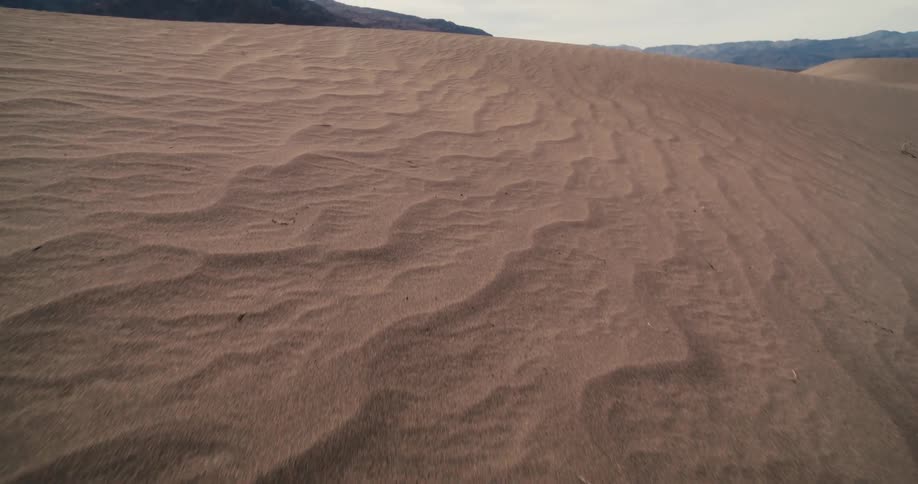 The sun shines on ripples of sand in the desert and there are mountains in the distance under a cloudy sky