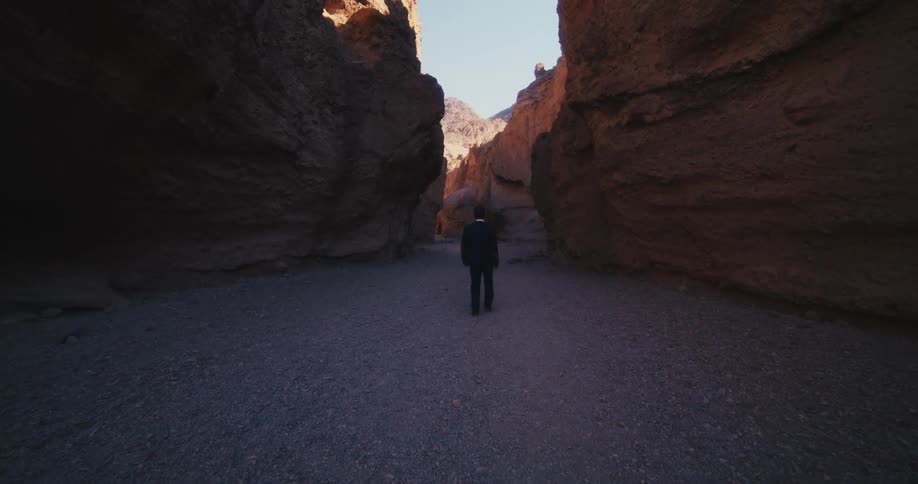A man in a suit walks along the floor of a canyon during the day