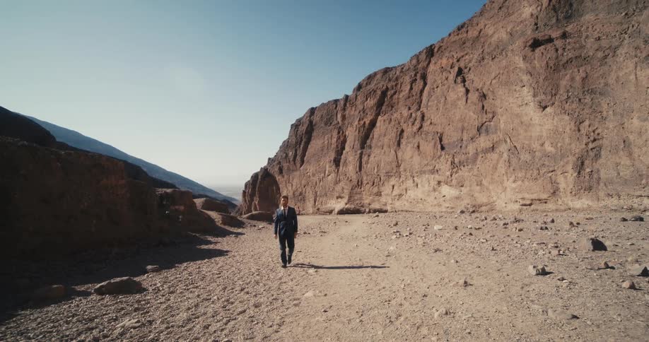 A man wearing a suit is walking in the desert badlands along a plateau with mountains in the distance