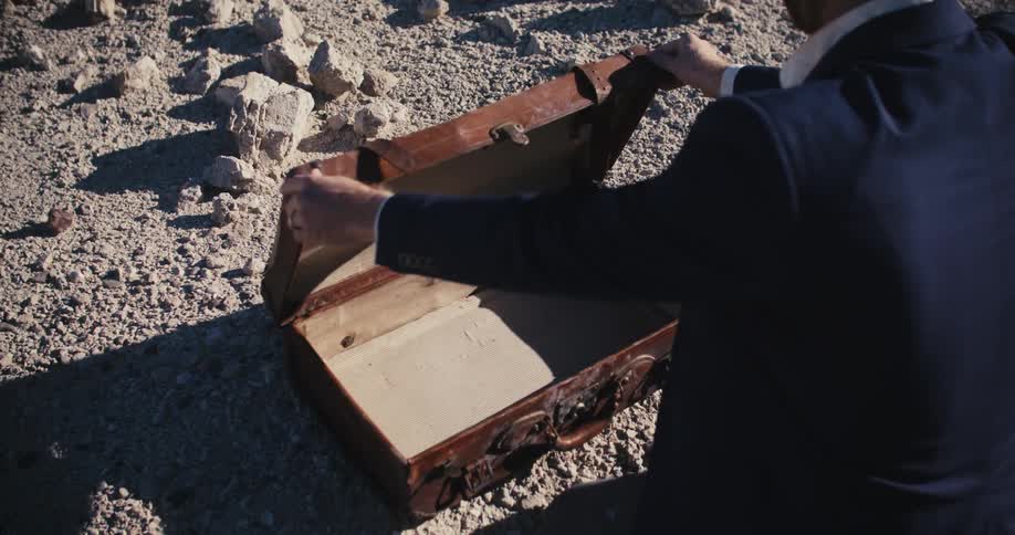 A man kneeling outdoors in gravel opens a suitcase only to find that it is empty