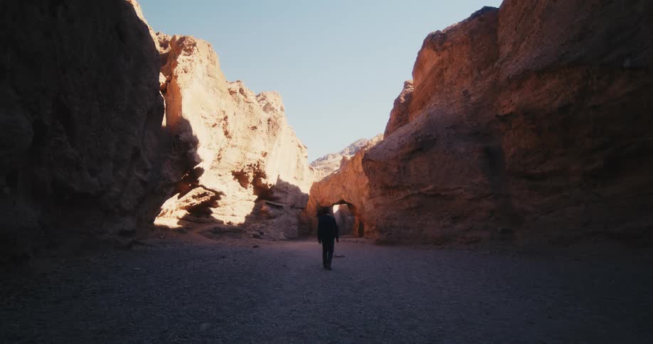 A man in a suit looks around at his surroundings while walking awkwardly along the floor of a canyon