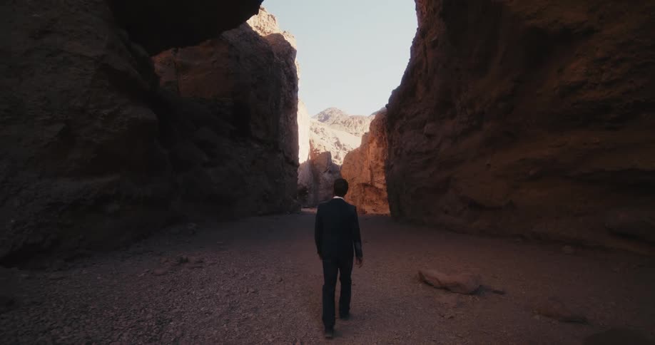A man in a suit walks beneath a natural arch in a canyon during the day