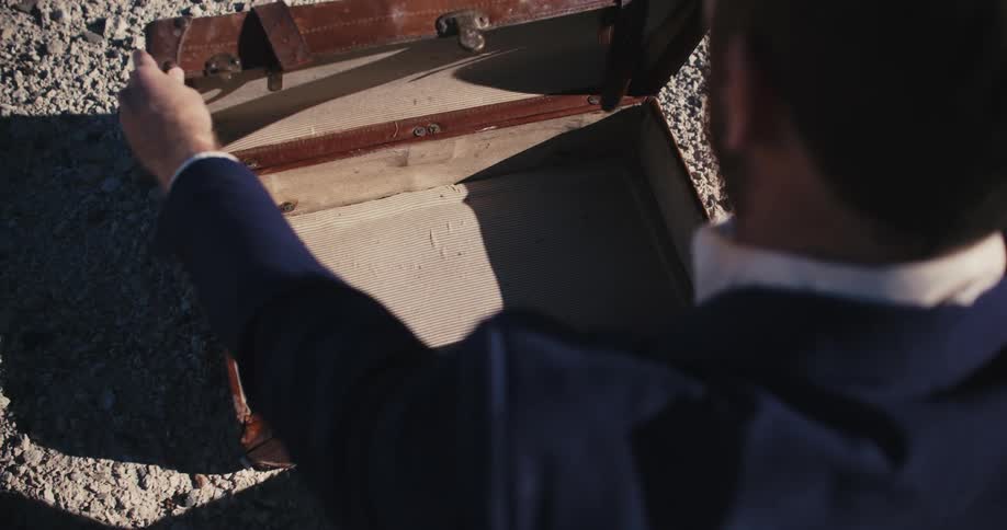 A man wearing a suit is sitting in the desert sand as he opens an empty briefcase and then closes it