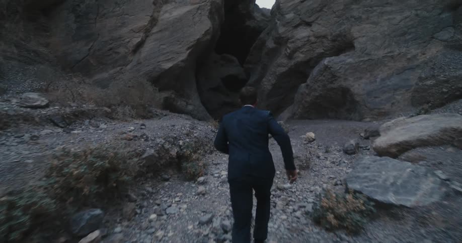 Man in a business suit walking then running towards rocks in the valley