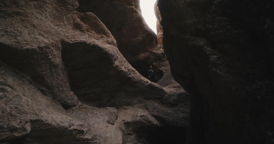 A man wearing a suit is standing outside of a mountain cave as light shines into the crevasse from above