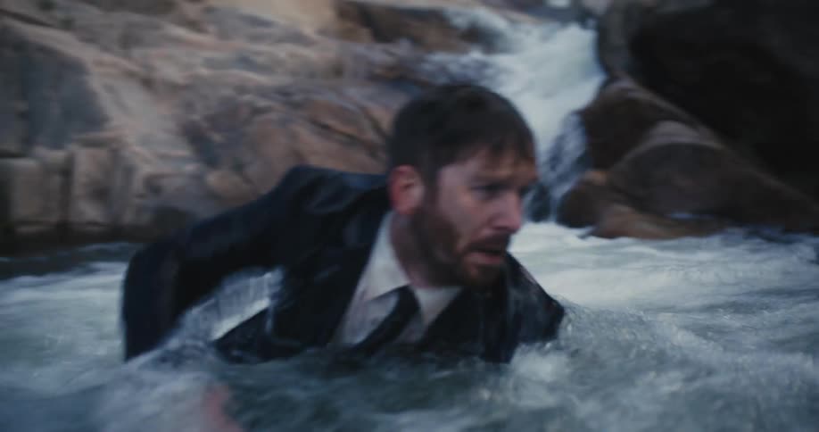 A man in a suit emerges from beneath the surface of a river and struggles to reach the shore