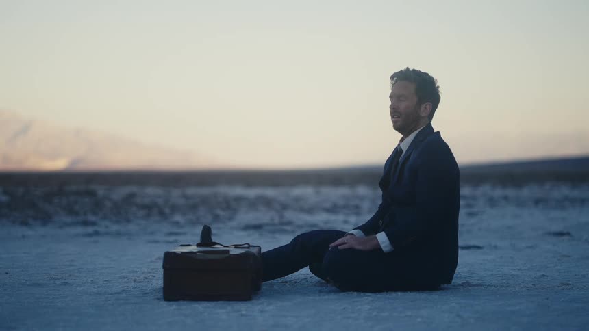 A distressed man in a suit sits in the dirt next to a briefcase in the desert during the day