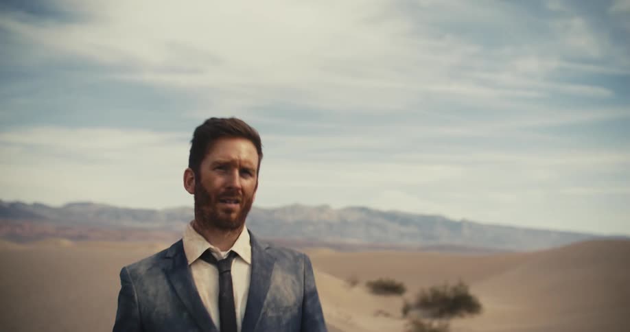 A man wearing a dirty suit appears to be tired and stumbling as he walks through the desert and there are mountains in the distance