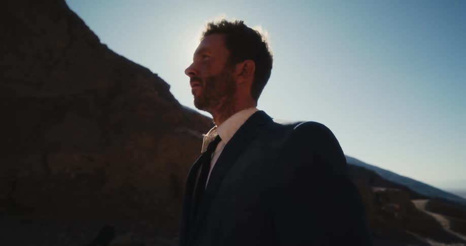 The sun shines over a mountain as a man in a suit walks across the desert