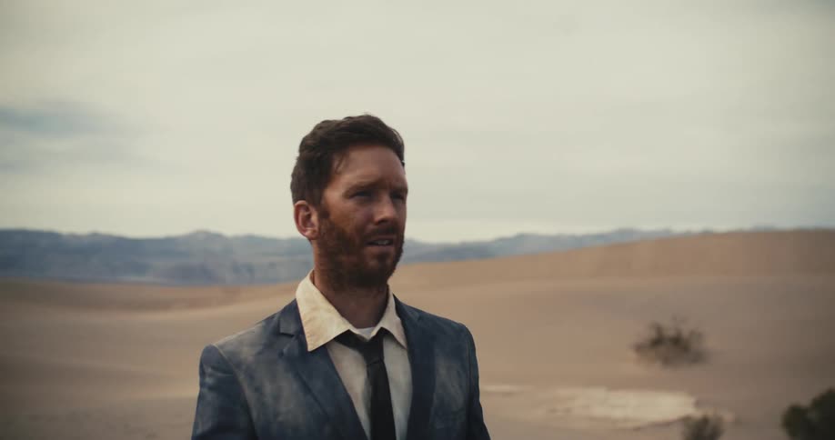 An exhausted man in a dirty suit slowly walks through a desert during the day