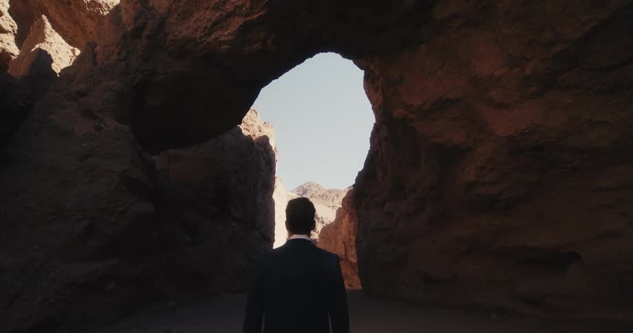A man in a suit stands in a canyon while looking up at a natural arch of rock above him