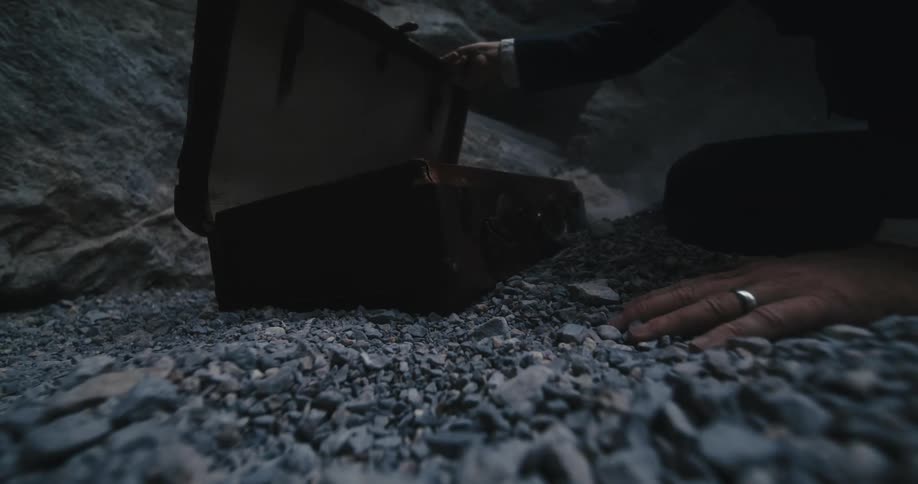 Man crashes onto his knees as he rushes to open case sitting on gravel near rock slope